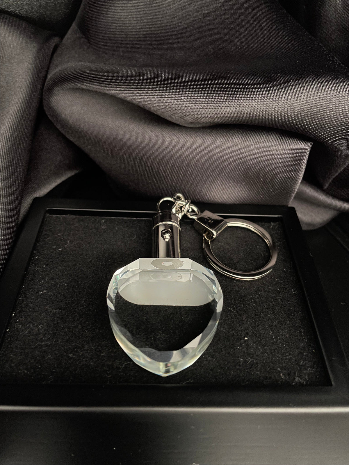 A clear faceted crystal keychain with a silver metal split ring and connector, displayed inside a black presentation box lined with black fabric next to draped dark gray satin.